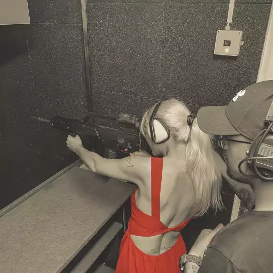 Blonde girl in red dress at a gun range in Budapest shooting a pistol under a supervision of an instructor.