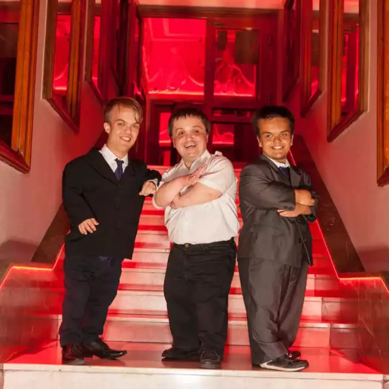 Three dwarf entertainers dressed up in suits posing on a staircase.